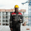 Mature African American builder with rolled blueprint looking at camera Mature African American builder with rolled blueprint looking at camera while standing against unfinished buildings at construction site